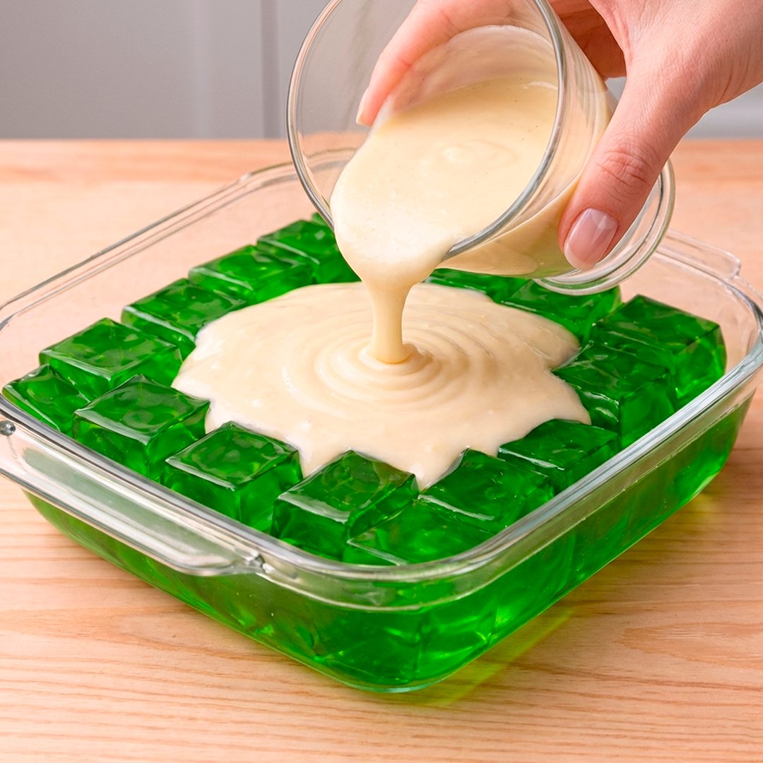 Hand pouring condensed milk cream over emerald green jelly cubes arranged in a glass dish on a wooden surface